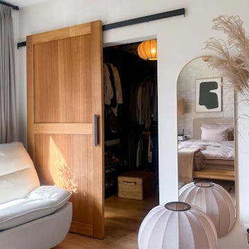 Offene Schiebetüren aus Eichenholz führen von einem hellen, beigen Schlafzimmer in einen praktischen begehbaren Kleiderschrank. Im Raum fallen ein bequemer Sessel und eine stimmungsvolle Beleuchtung ins Auge.