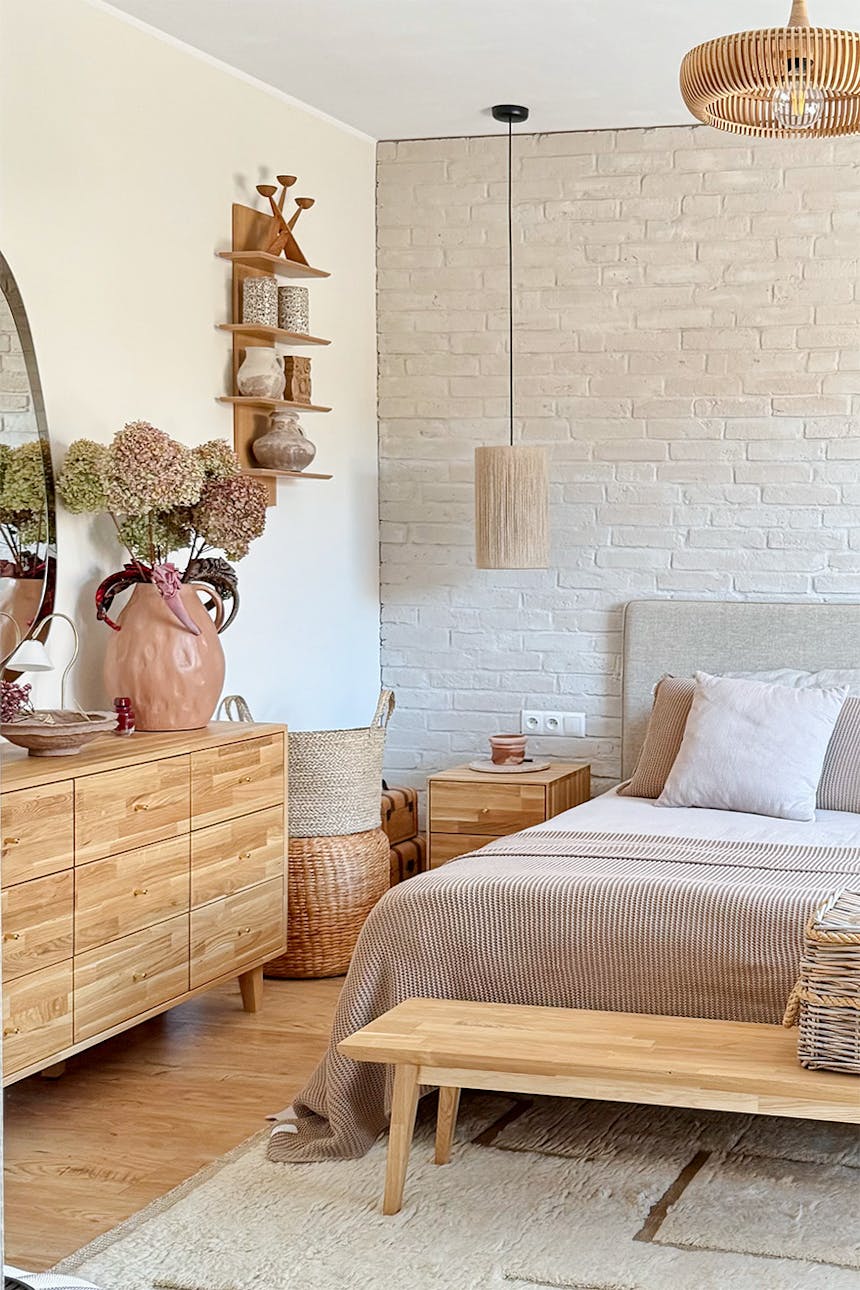 Ein Schlafzimmer mit einer Kommode aus Holz, einem Spiegel und natürlichen Dekoelementen. Zu sehen sind beigefarbene Tagesdecken, geflochtene Körbe und getrocknete Pflanzen in einer Vase.