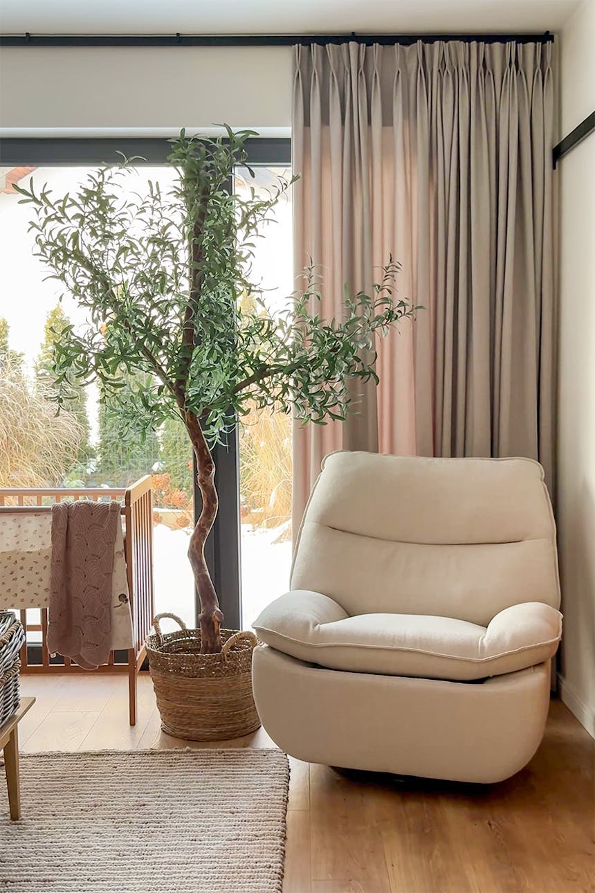 Ein heller Entspannungsbereich im Schlafzimmer mit einem cremefarbenen Sessel und einem großen Olivenbaum in einem geflochtenen Korb.