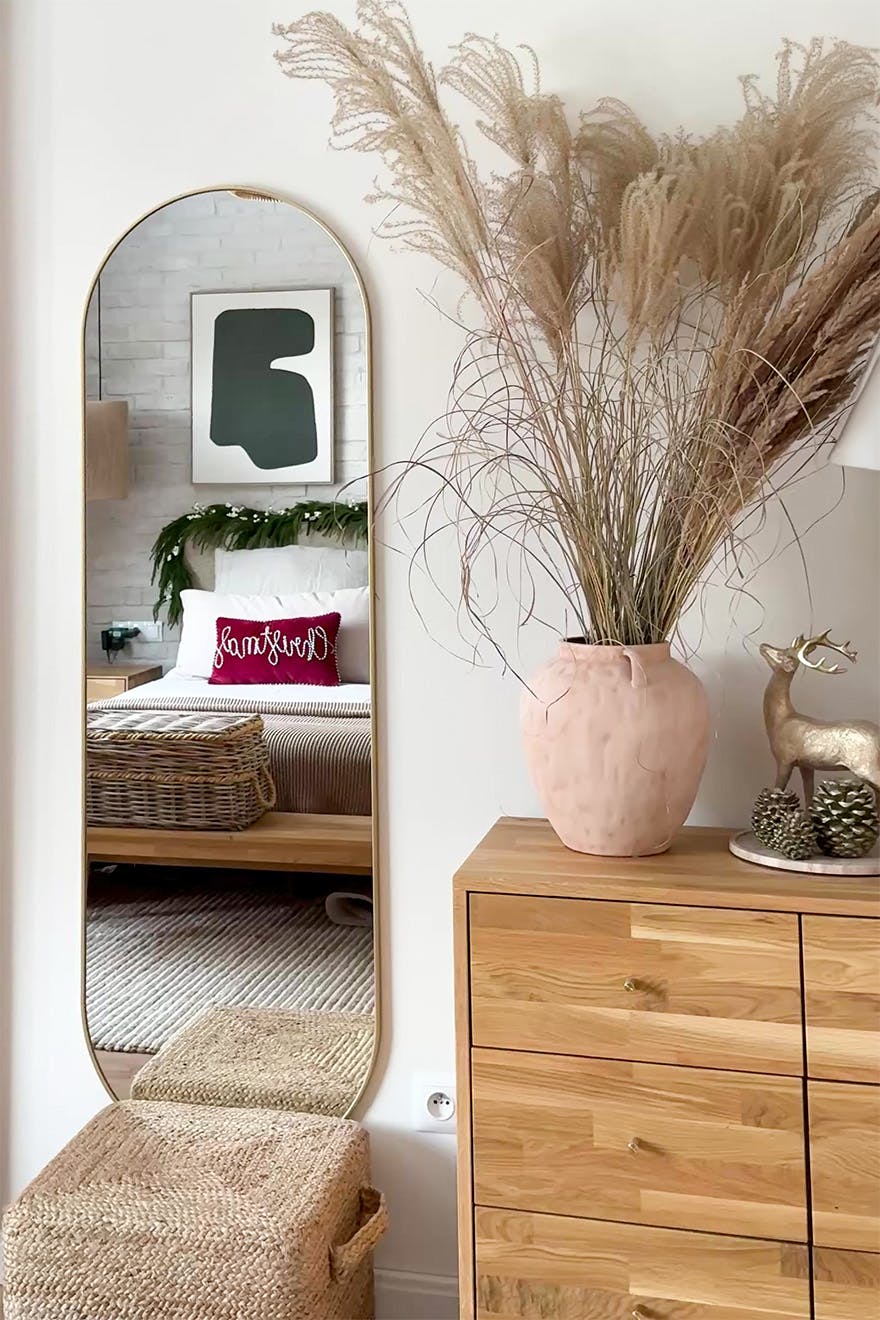 A wooden chest of drawers with a decorative vase and dried grasses. Next to it stands a tall, curved mirror, in which the bed in the bedroom is reflected.