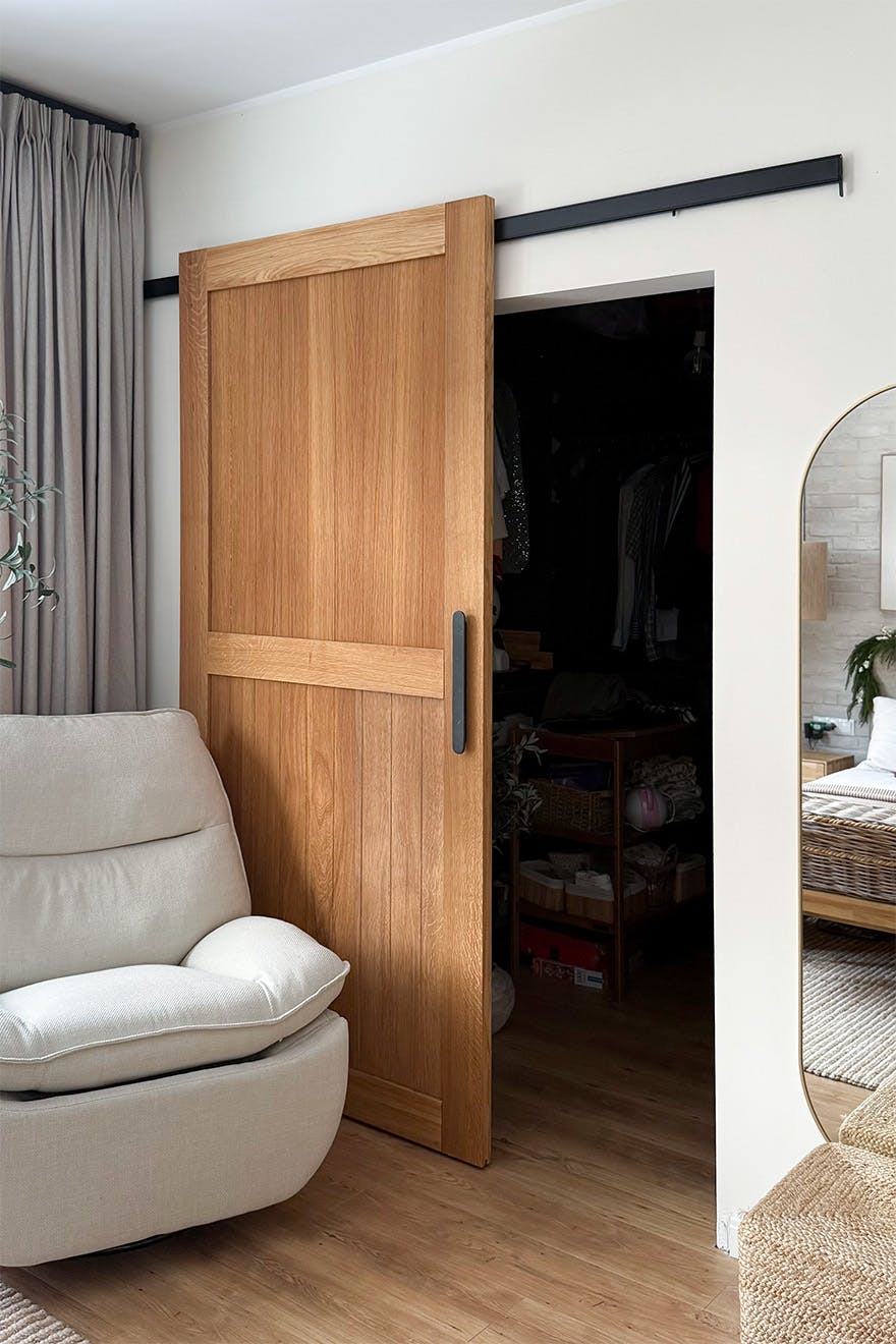 A wide shot of the oak sliding doors revealing the interior of the walk-in wardrobe. The room features a beige armchair, light-coloured curtains and a section of a mirror reflecting the bedroom.