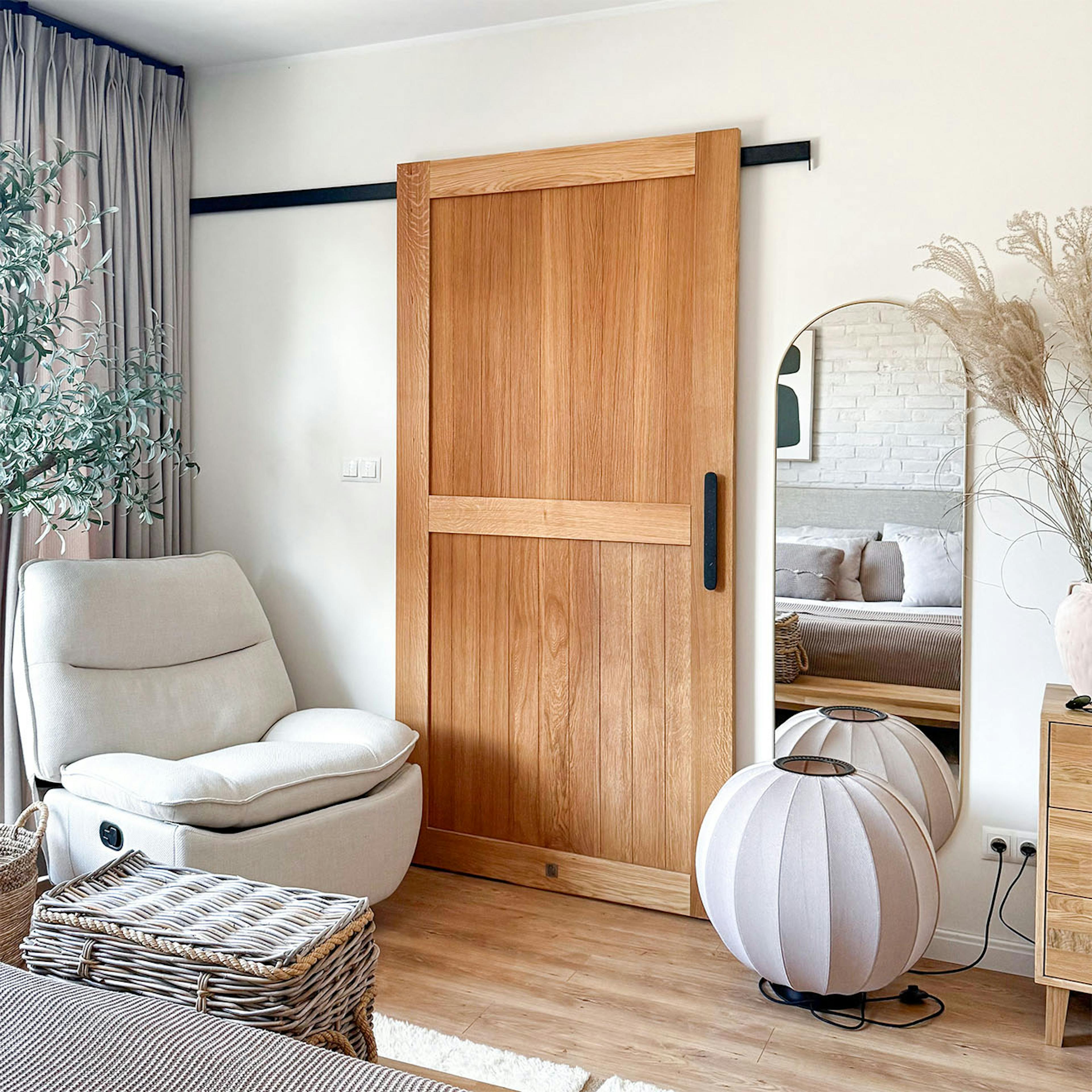 Une porte coulissante en chêne fermée, guidée par un rail noir, dans une chambre à coucher chaleureuse. À côté, un grand miroir, des accessoires tressés et des textiles clairs dans le style « slow living ».