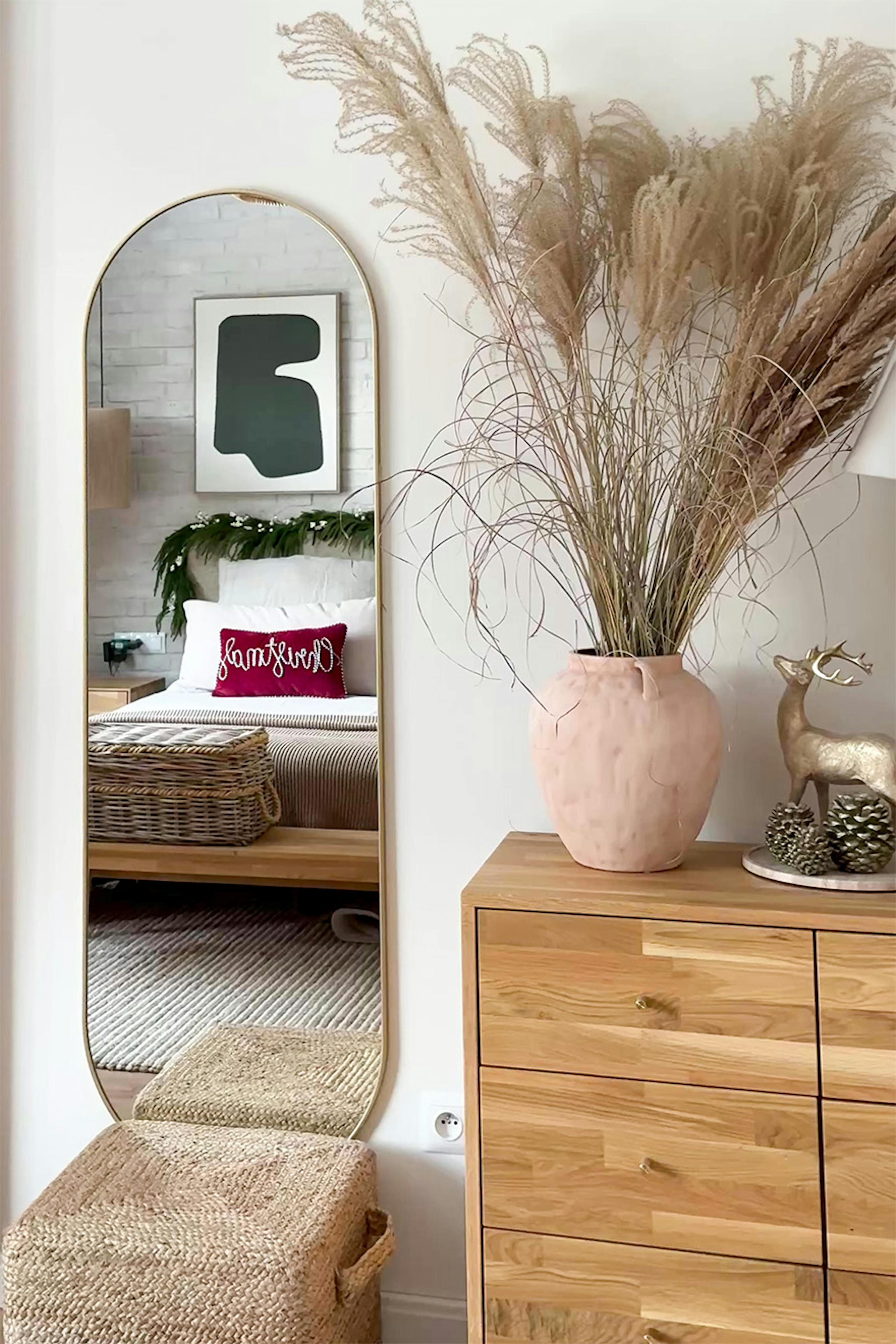 Une commode en bois ornée d'un vase décoratif et d'herbes séchées. À côté, un grand miroir aux contours arrondis dans lequel se reflète le lit de la chambre.