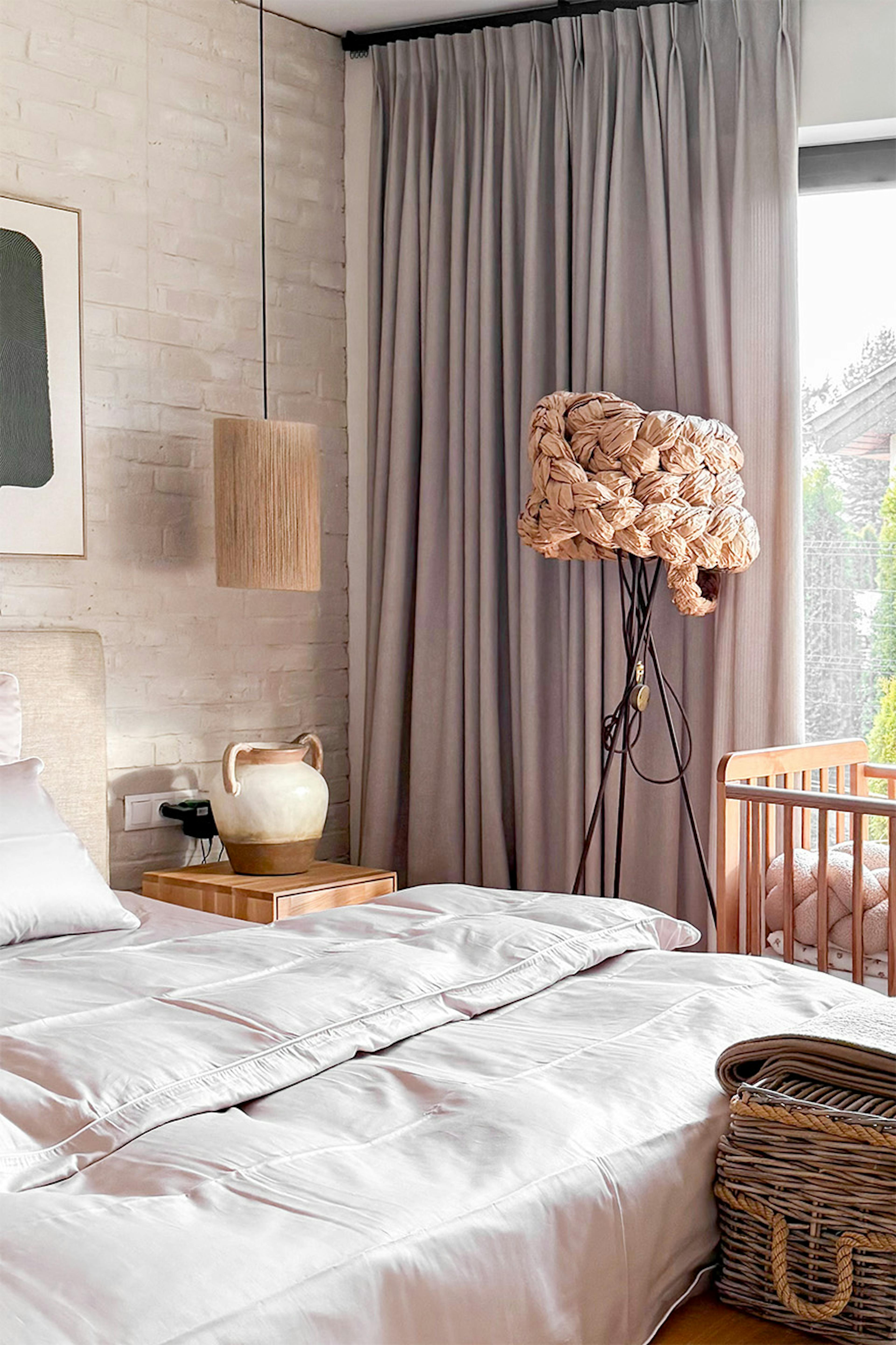 Un coin douillet dans la chambre à coucher, avec un lit bébé, un rideau en lin et une lampe d'ambiance aux formes organiques. En arrière-plan, un mur en briques blanches.