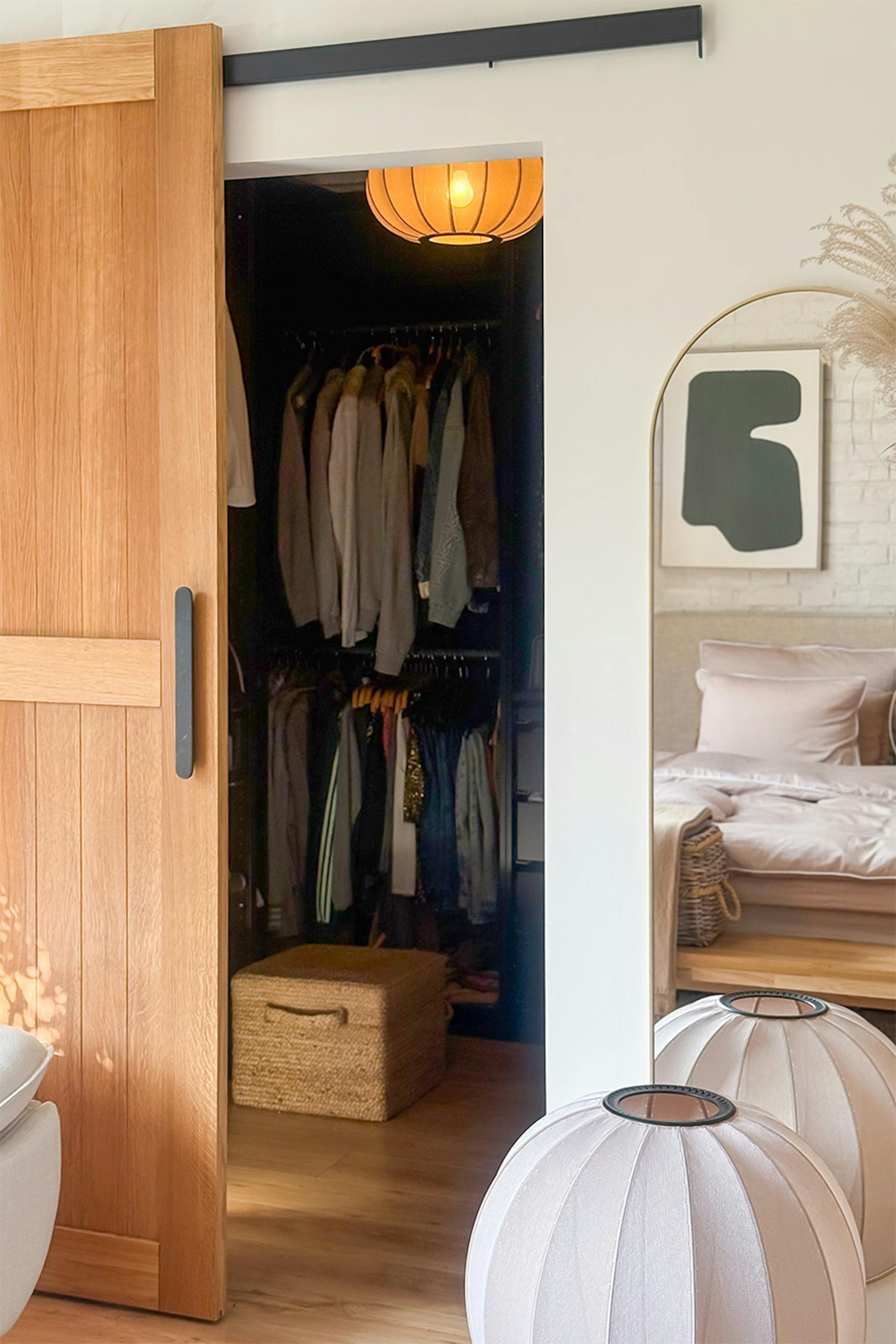 On accède au dressing par une porte coulissante en chêne. À l'intérieur, on aperçoit des vêtements bien rangés, ainsi qu'un miroir et des lampadaires design.