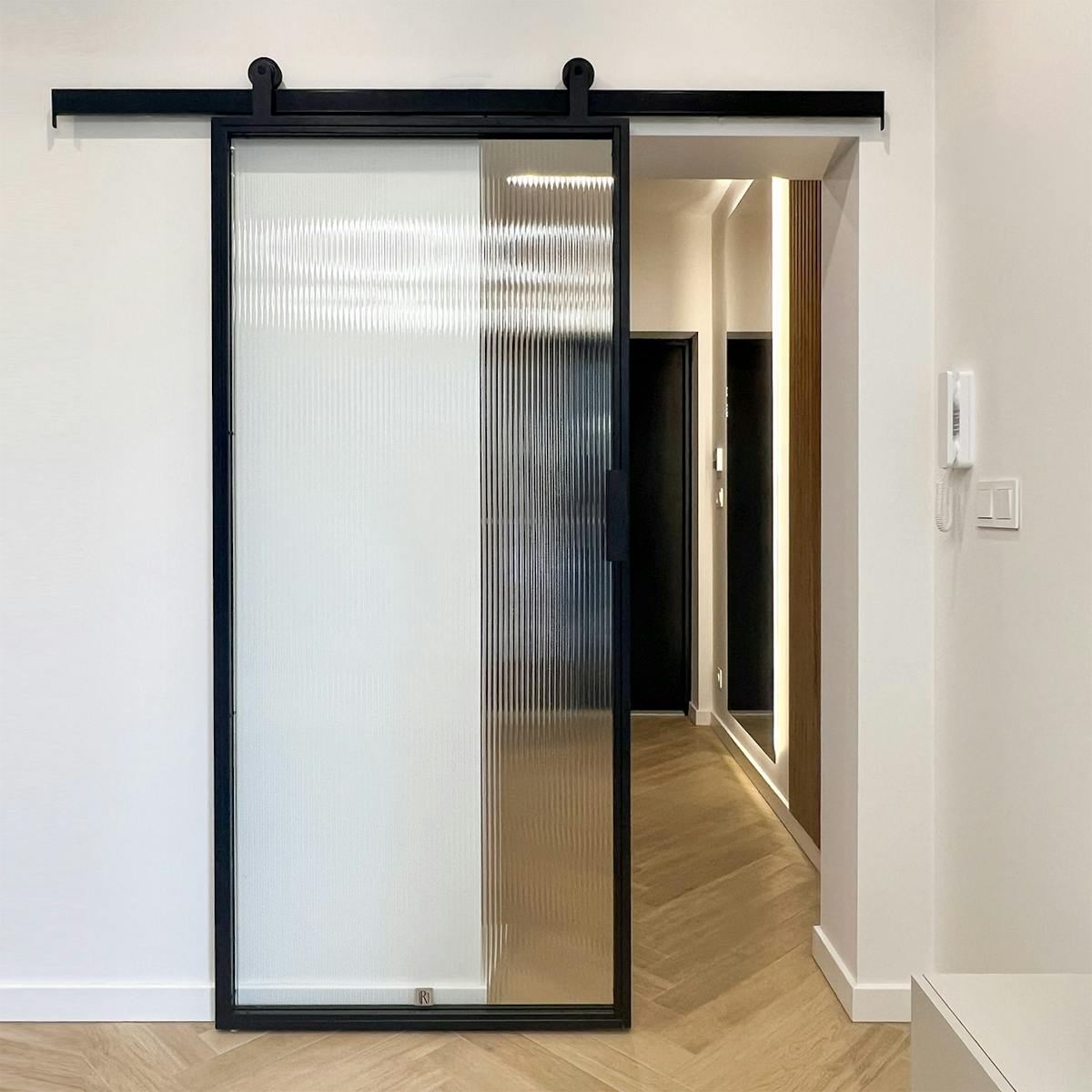 Czarne drzwi przesuwne ze szkłem ryflowanym, widok na przedpokój z drewnianymi lamelami i nowoczesnym oświetleniem.