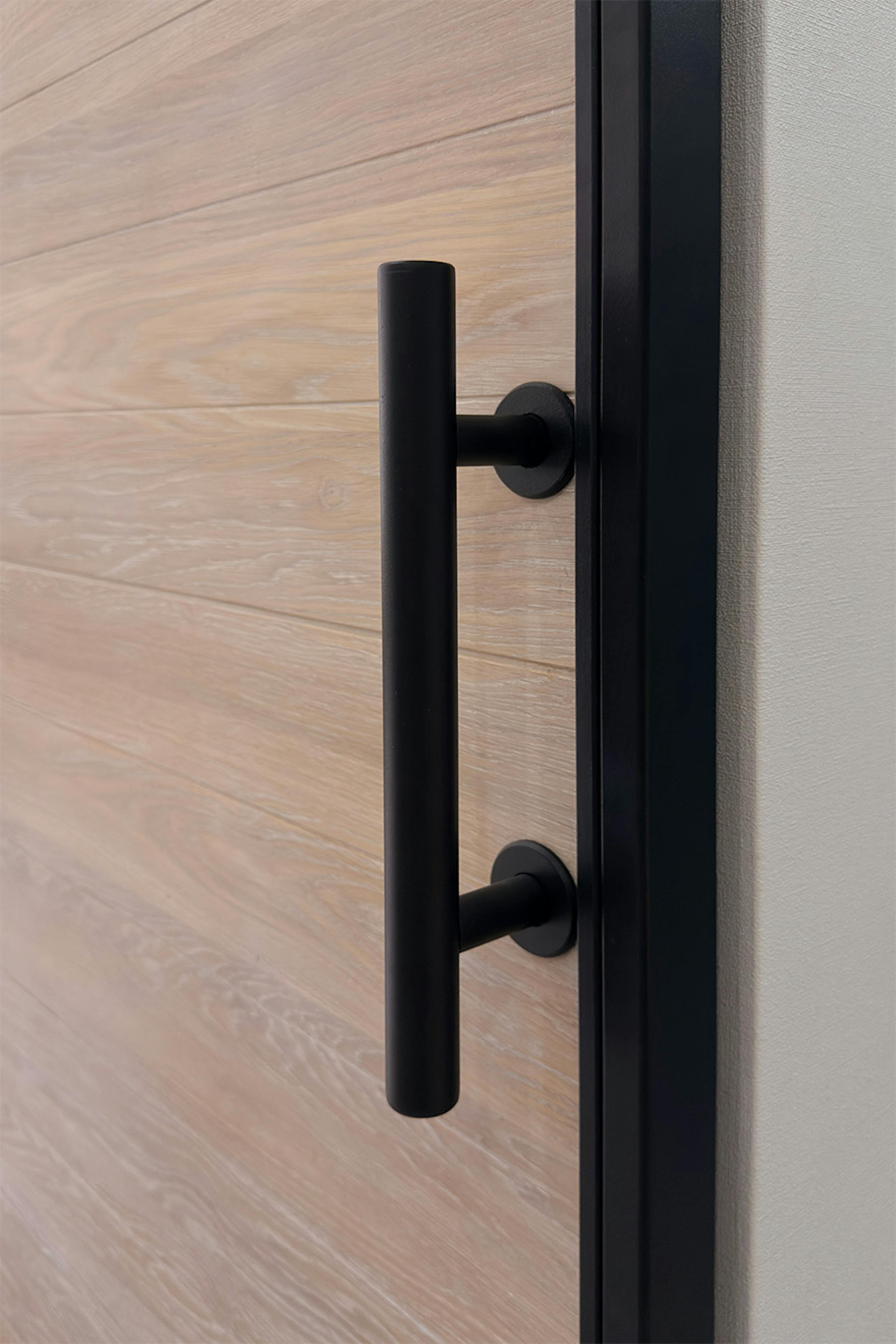 Gros plan sur une poignée verticale noir mat fixée sur un panneau de porte coulissante en chêne. La texture et le grain du bois ressortent clairement grâce à la finition en chêne blanchi.