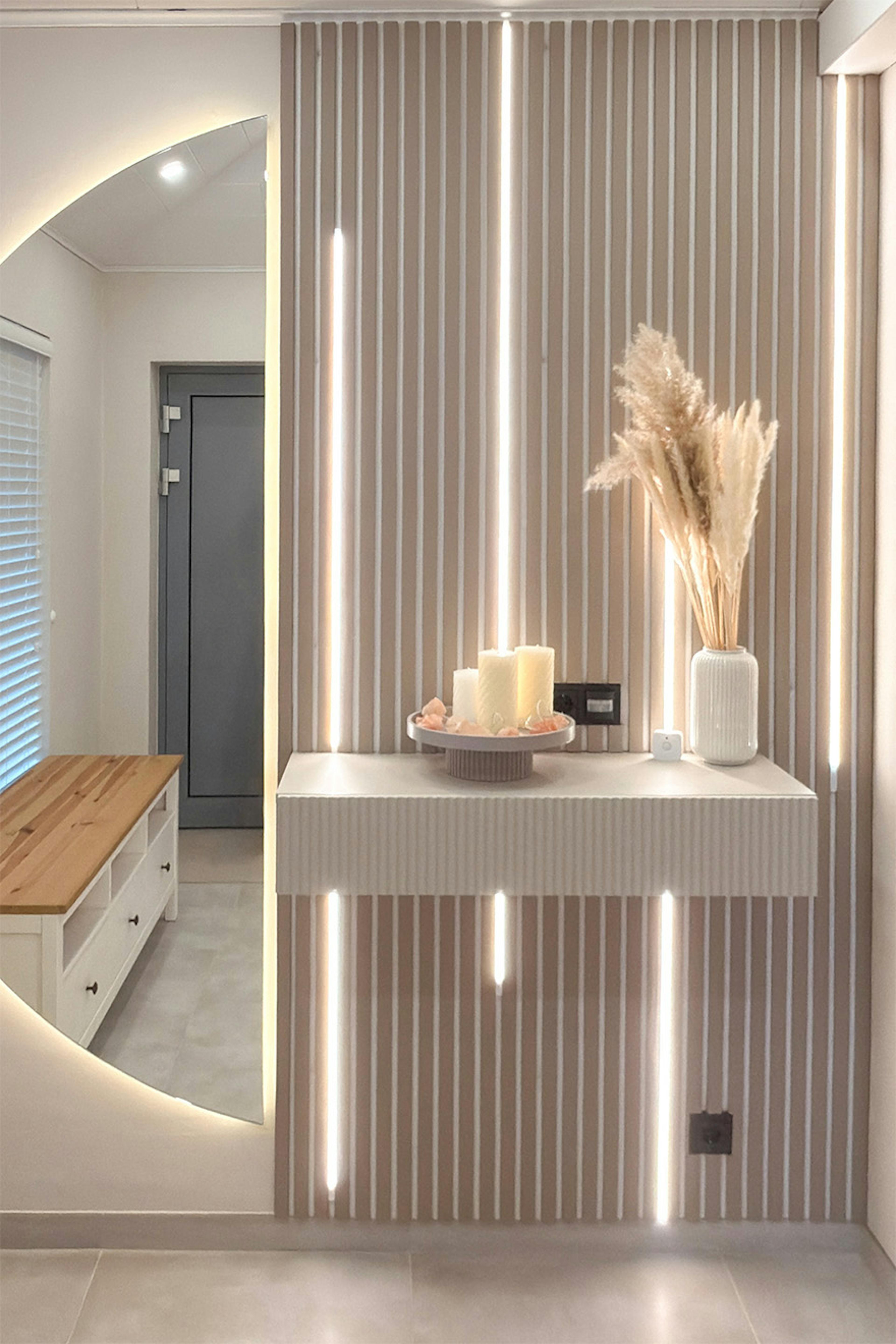 Un mur décoratif composé de lattes verticales et de bandes LED intégrées dans un couloir lumineux. Une console murale accueille des bougies décoratives et un vase rempli d'herbe de la pampa. Un grand miroir rond rétroéclairé est accroché au mur juste à côté.