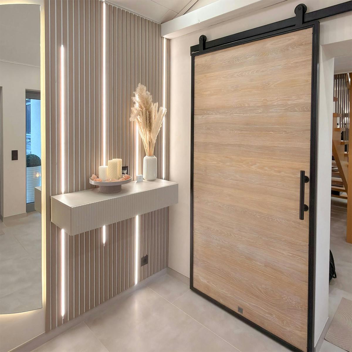 Nowoczesna strefa wejściowa w stylu warm minimalism. Na ścianie dekoracyjne lamele z pionowym podświetleniem LED i wisząca konsola, obok dębowe drzwi przesuwne w loftowym stylu