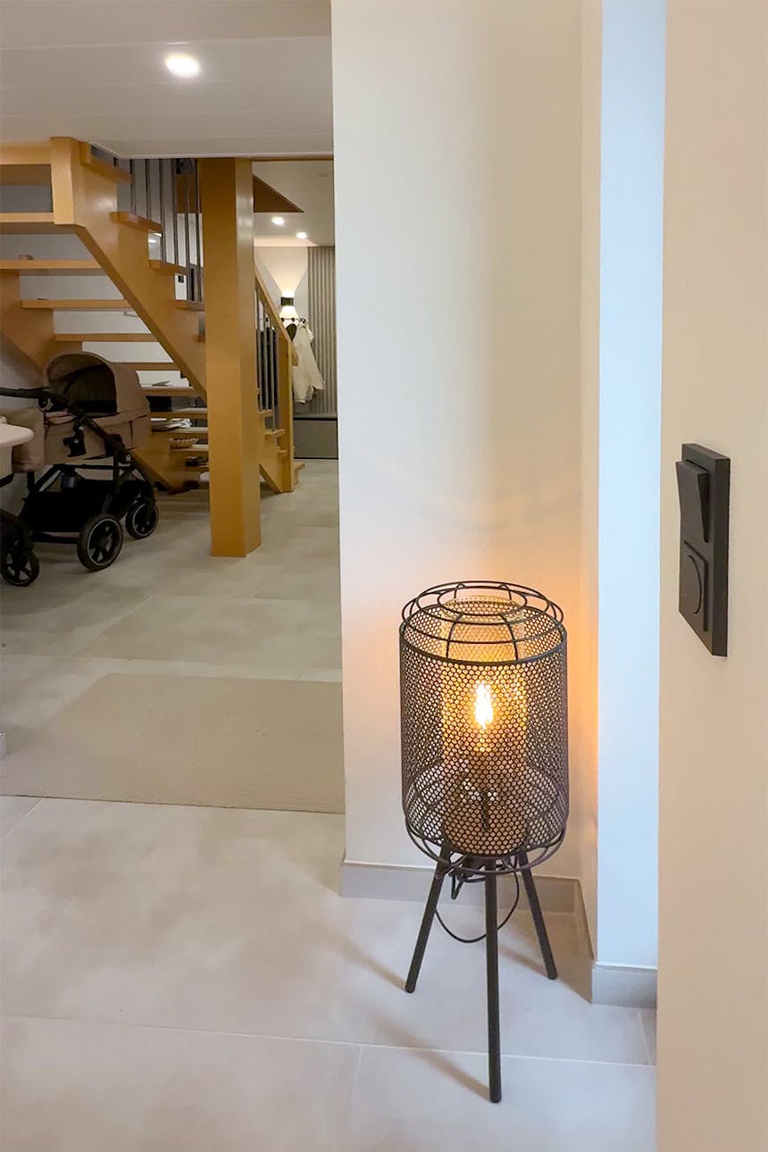 Czarna, stojąca lampa ażurowa z metalowej siatki typu tripod, rzucająca ciepłe światło w rogu pomieszczenia.