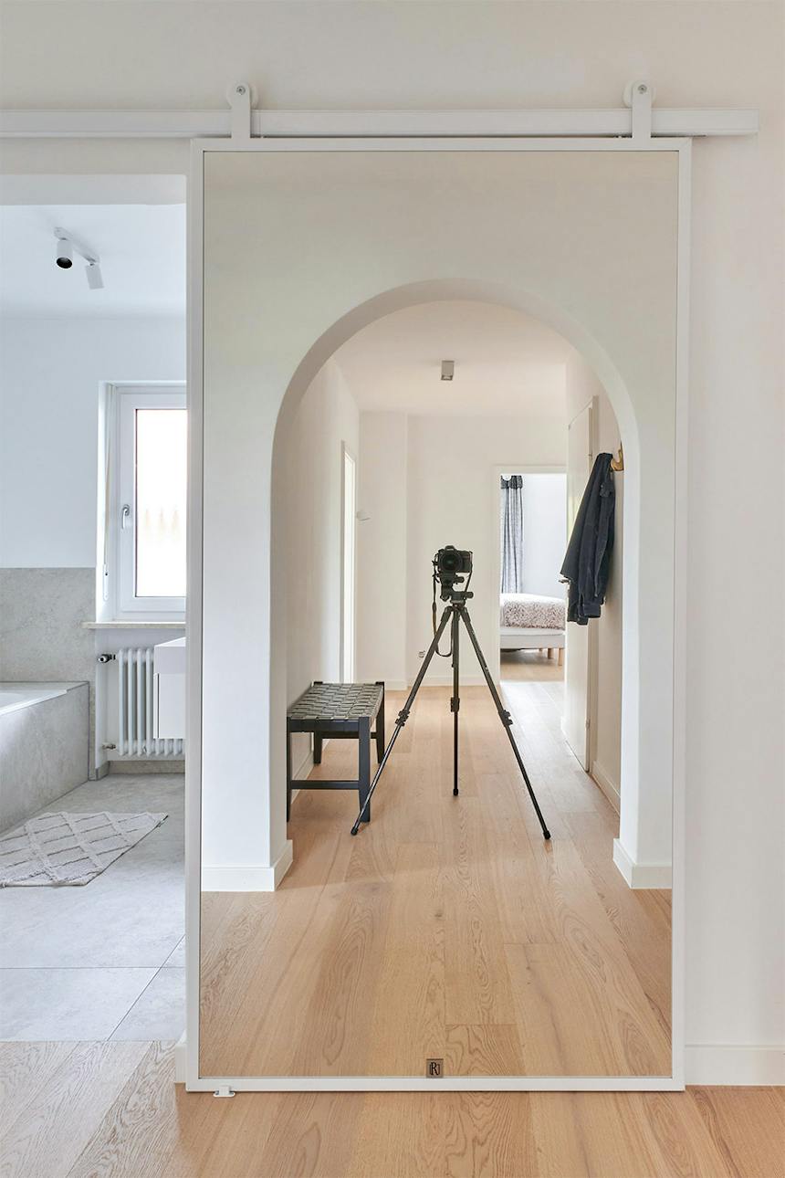 Weiße Spiegelschiebetüren am Eingang zum hellen Badezimmer, in denen sich der Flur mit seinem architektonischen Rundbogen und dem Eichenparkett widerspiegelt.