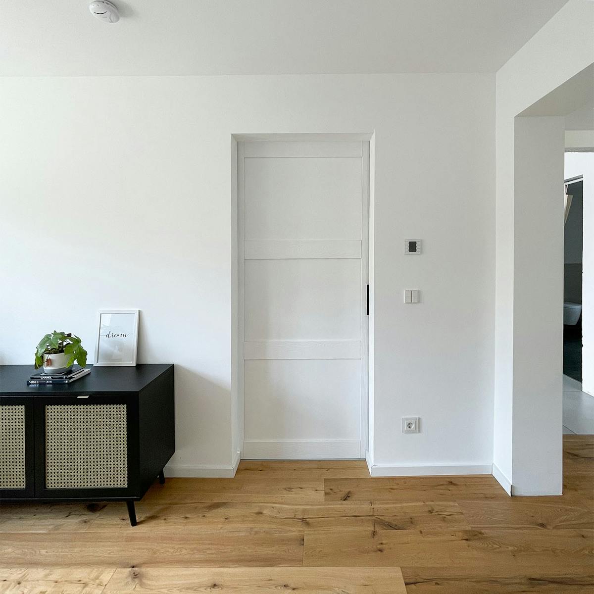 Schwarze niedrige Kommode mit Rattan auf weißem Wandhintergrund, weiße Massivholz-Schiebetür, weiße Tür auf weißer Wand