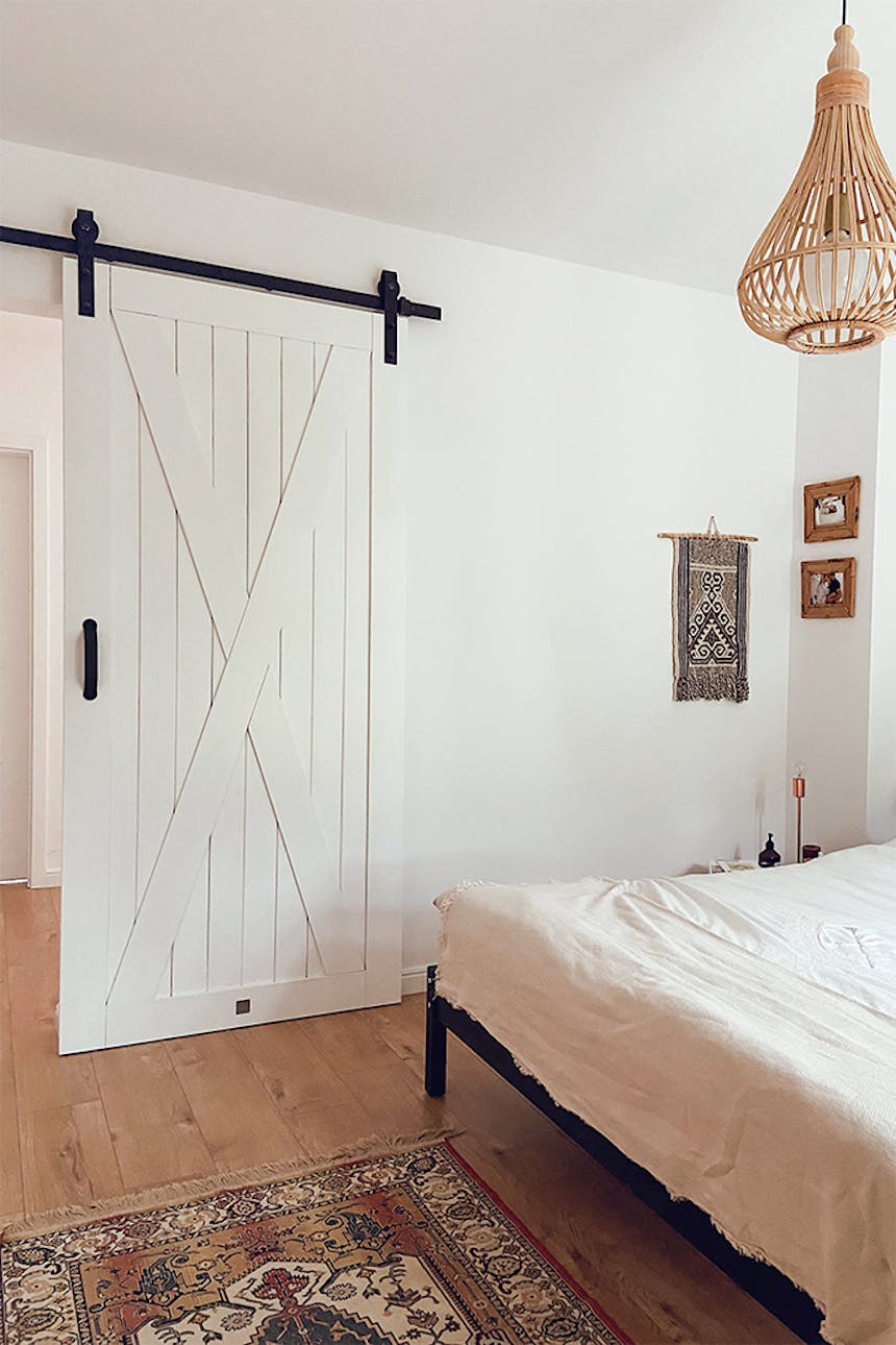 Weiße Massivholz-Schiebetür im Eingang zum Naturschlafzimmer, bunter Teppich vor dem Bett im Schlafzimmer, weiße Wände im Schlafzimmer, Bambus-Pendelleuchte, natürliche Boho-Dekorationen an der weißen Wand