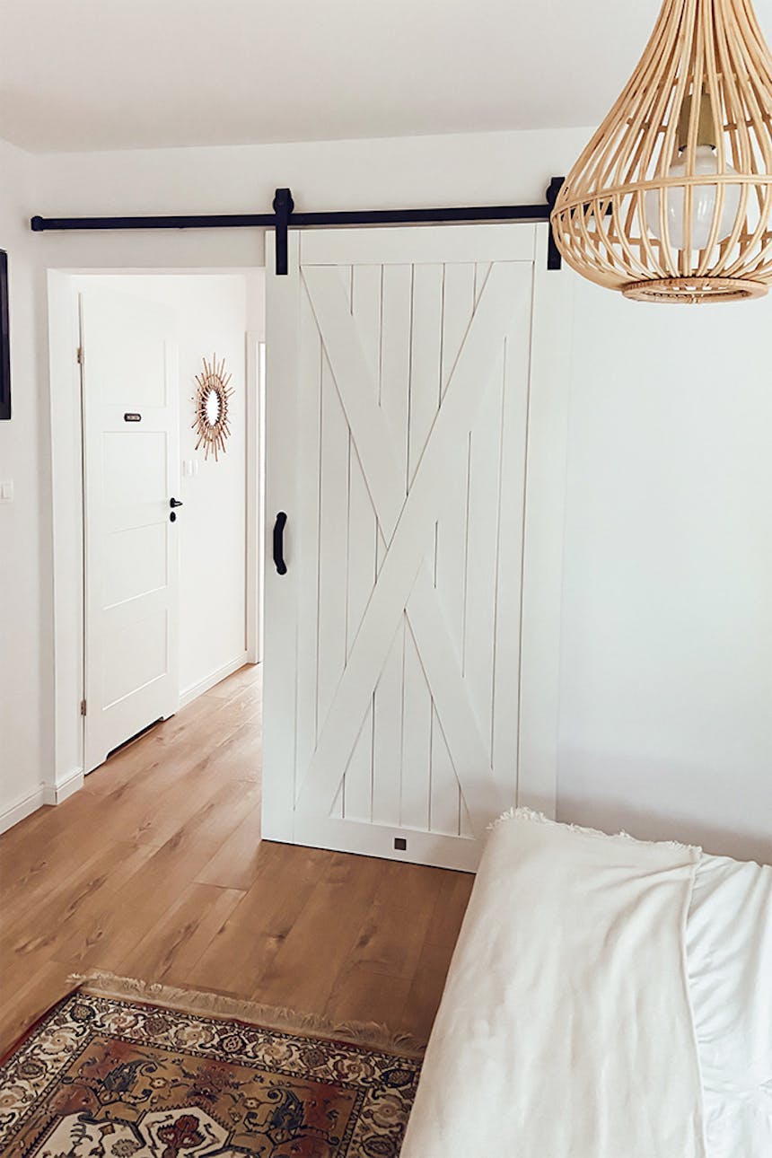white sliding door in the entrance to the bedroom, white walls and wooden floor in a natural bedroom, bamboo lampshade, colourful, decorative rug in front of the bed