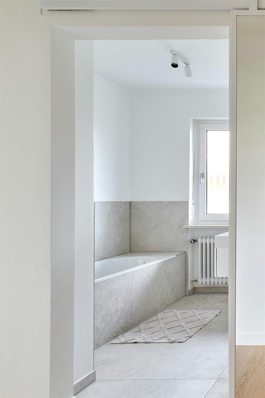 Offene Schiebetüren mit Spiegel, die den Eingang zu einem minimalistischen Badezimmer mit Badewanne und grauen Fliesen freigeben.