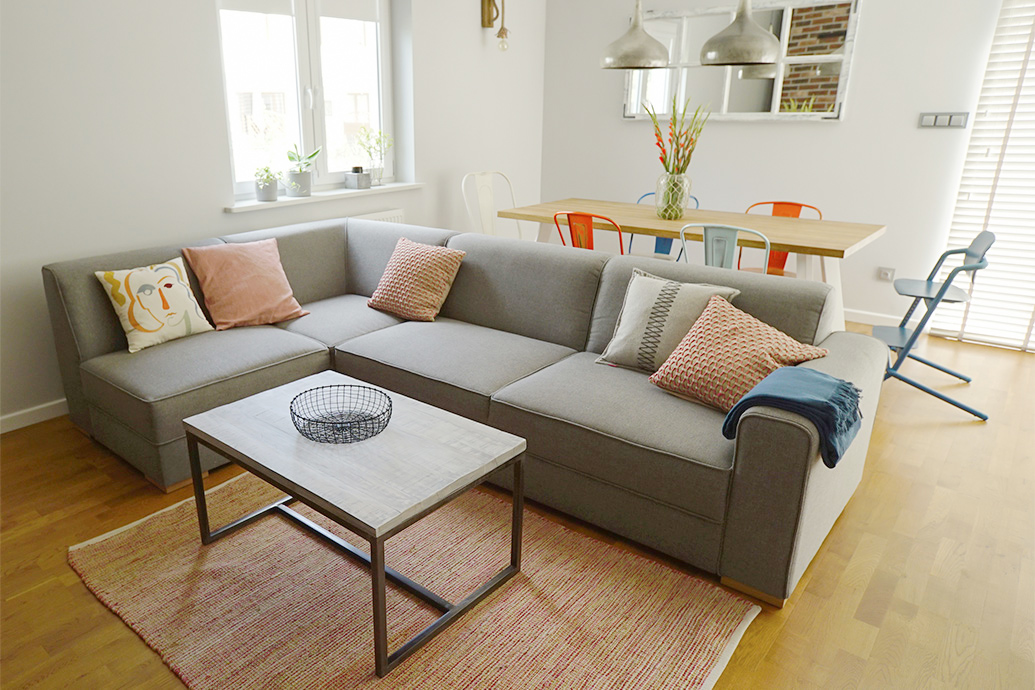 großes graues Sofa, dekorative Kissen, Couchtisch auf Metallbeinen, bunte Stühle im Esszimmer