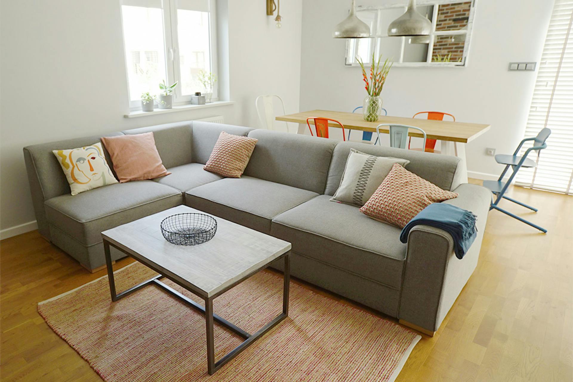 großes graues Sofa, dekorative Kissen, Couchtisch auf Metallbeinen, bunte Stühle im Esszimmer