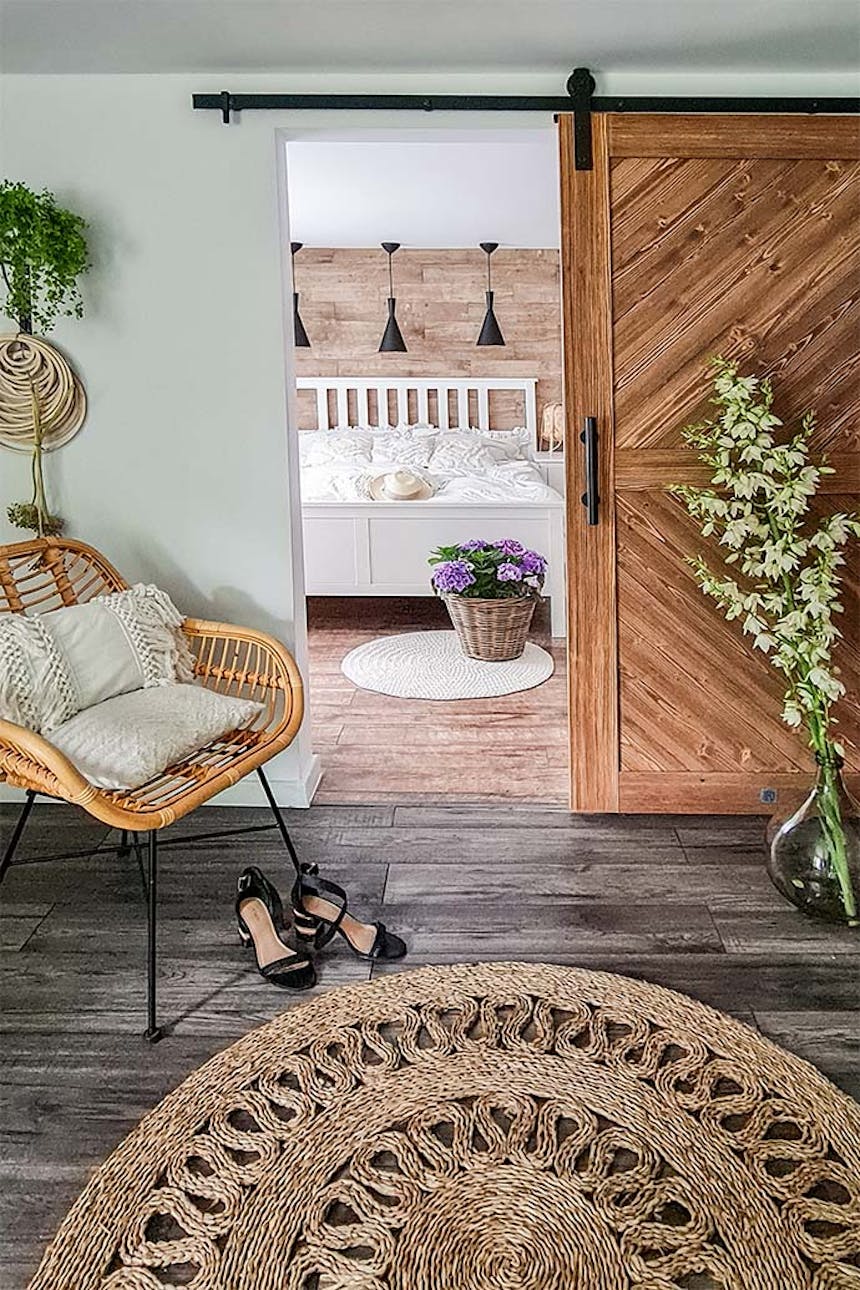 Holzschiebetür im Schlafzimmereingang, Korbsessel mit dekorativen Kissen, geflochtener runder Juteteppich, Blumenschmuck in einem Weidenkorb, weißes Bett in einem Schlafzimmer in skandinavischem Interieur, schwarze Pendelleuchten über einem weißen Bett