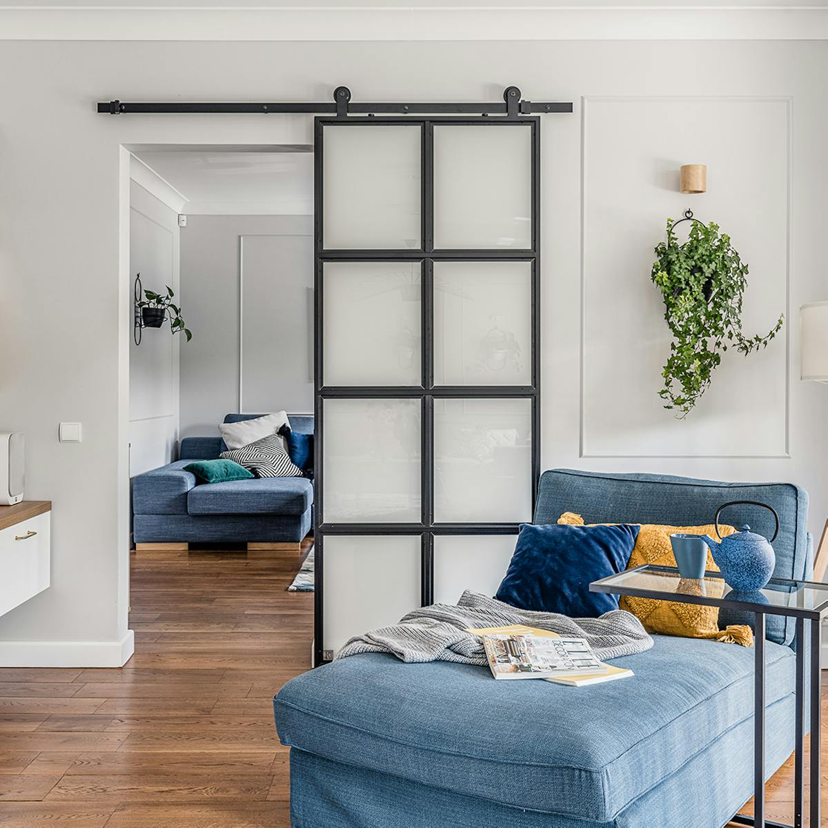 Leseecke, blaues Denim Sofa, Metall- und Glasschiebetür, Industrietür, heller Raum, blaue und gelbe dekorative Kissen, Wandblumenbeet  