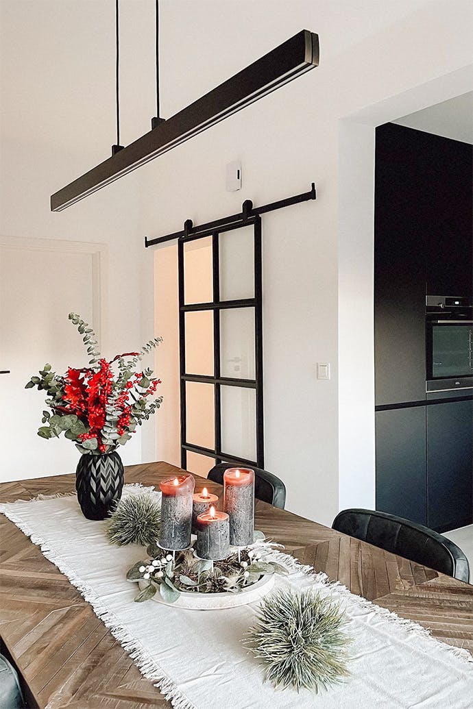 Des bougies décoratives, un vase de fleurs séchées sur une table à manger en bois éclairée par une longue lampe minimaliste