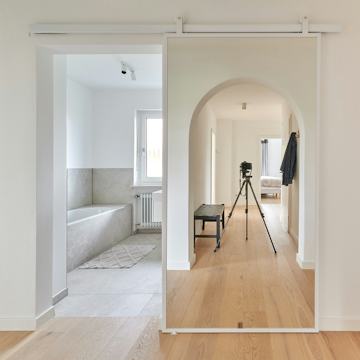 Weiße Spiegelschiebetüren am Eingang zum hellen Badezimmer, in denen sich der Flur mit seinem architektonischen Rundbogen und dem Eichenparkett widerspiegelt.