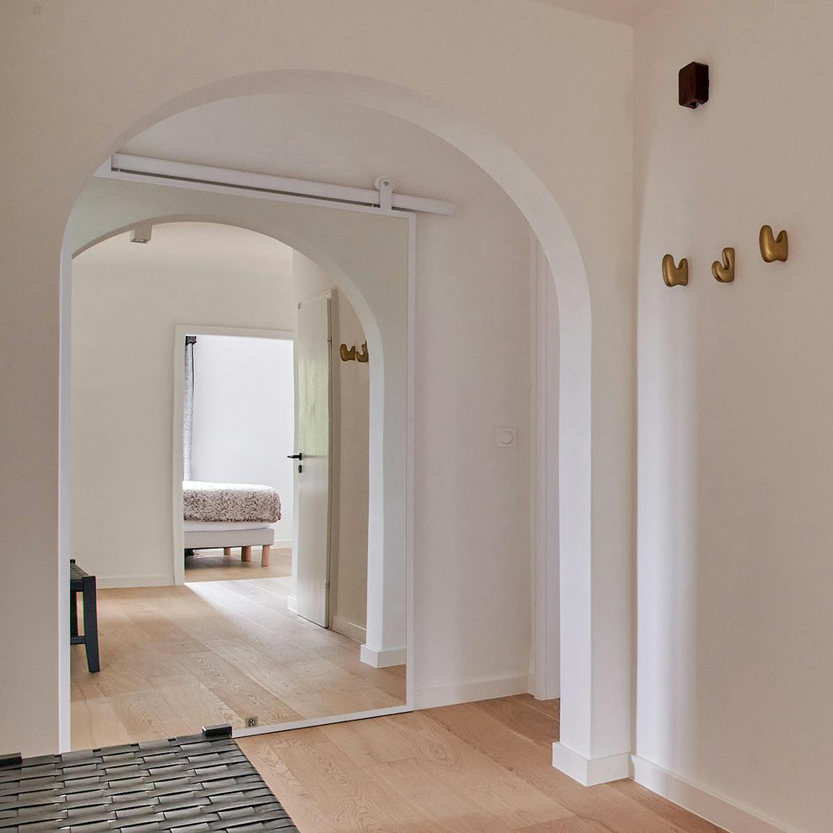 Der Blick durch den bogenförmigen Durchgang auf die weißen Schiebetüren mit Spiegel, die den Raum der Wohnung widerspiegeln, sowie auf die Messingkleiderhaken an der Wand.