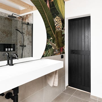 Schwarze Holzschiebetüren, gesehen vom Inneren eines modernen Badezimmers mit schräger Decke, weißem Waschbecken und einer Tapete mit tropischem Motiv.